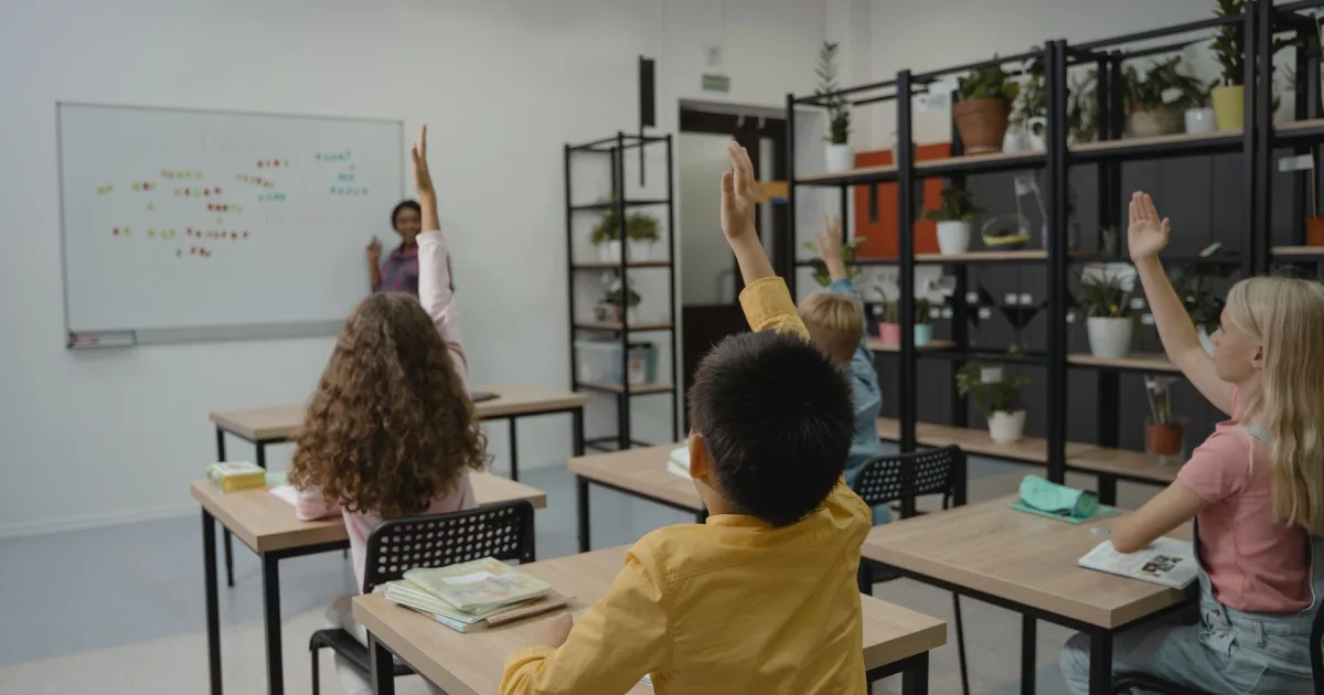 Students raising their hands in a classroom with a teacher standing by a whiteboard.