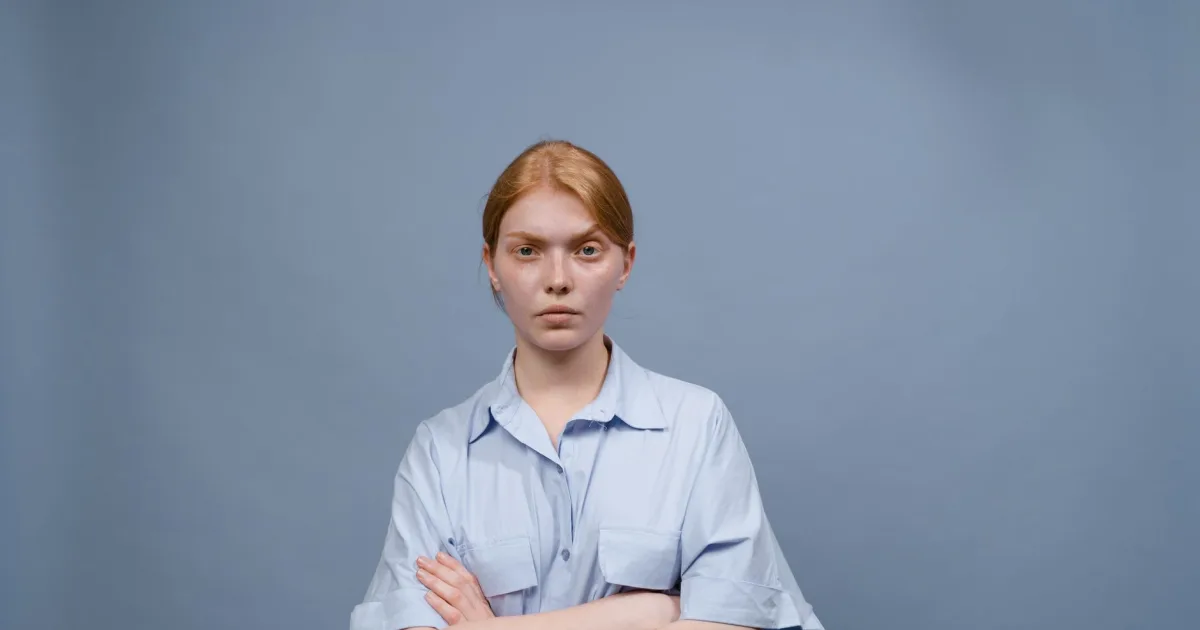 Red-haired woman in a light blue button-down shirt, arms crossed, against a plain blue background.